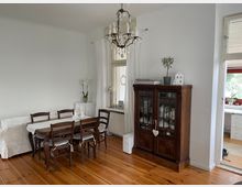 Esszimmer mit Holzboden, einem großen Holztisch und sechs Stühlen. Im Hintergrund stehen eine Vitrine aus dunklem Holz, ein kleines weißes Eckmöbel, weiße Vorhänge an großen Fenstern und eine dekorative Hängelampe. Rechts führt eine offene Tür in einen angrenzenden Raum mit Blick ins Freie.