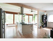 Geräumige, moderne Wohnküche mit heller Holzoptik und großen Fenstern, die Blick auf eine grüne Außenlandschaft bieten. Zentral steht eine Kücheninsel mit Herd und Dunstabzugshaube, umgeben von Pflanzen, einem Bücherregal und Sitzgelegenheiten.