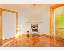 Wohnraum mit Kamin an einer hellen Wand, verziert mit feinem Ornamenttapetenmuster. Der Boden besteht aus glänzendem Parkett, und der Raum hat Türen mit gelben Holzrahmen, von denen eine in ein Zimmer mit grüner Wandfarbe führt.