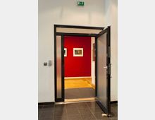 Eine teilweise geöffnete Glastür führt in einen Innenraum mit roter Wand, an der zwei gerahmte Bilder hängen. Der Boden ist aus Holzdielen, und über der Tür befindet sich ein Notausgangsschild.