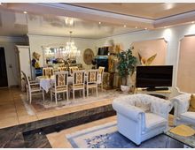 Wohnzimmer mit luxuriöser Einrichtung: Ein Essbereich mit einem Tisch und zwölf verzierten Stühlen steht unter einem Kronleuchter, der Raum ist dekoriert mit goldfarbenen Statuen, einem Spiegel, Pflanzen und einem Fernseher. Die Wände sind in Weiß und Beige gestaltet, mit Paneelen und kunstvollen Verzierungen, auf dem Boden liegen Teppiche.