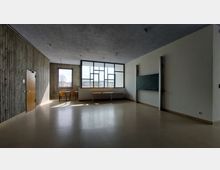 Klassenzimmer mit hellen Wänden und Betonakzenten, großen Fenstern mit geometrischem Rahmen, einer Wandtafel, einem Holzfensterrahmen und einfachen Möbeln wie Stühlen und einem Tisch. Der Raum hat einen minimalistischen Stil und ist von Tageslicht durchflutet.