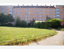 Eine mehrstöckige, pfirsichfarbene Wohnhausfassade mit weißen Fenstern, flankiert von Balkonen, steht im Hintergrund eines Parks mit einer großen Rasenfläche, umgeben von Bäumen, Sträuchern und einem geschwungenen Gehweg im Vordergrund.