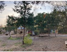 Ein Freigelände inmitten eines Waldgebietes mit mehreren einfachen, aus Holzplanken gebauten Hütten und Strukturen. Der Boden ist mit Erde und Laub bedeckt, umgeben von hohen Bäumen und vereinzeltem Unterholz.