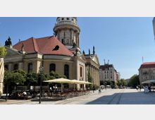 Historischer Platz mit einer großen Barockkirche mit rotem Dach und Säulen, flankiert von Statuen und Bäumen. Im Vordergrund stehen Tische und Stühle mit Sonnenschirmen, umgeben von gepflastertem Boden und weiteren Gebäuden im Hintergrund.