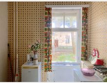 Kleine Küche mit Retro-Charakter, geprägt von gelb-gemusterter Tapete und einem großen Fenster mit Vorhängen im Vintage-Stil. Auf einer weißen Kühltruhe stehen eine Vase mit Kunstblumen und weitere Küchenutensilien, während ein Blick nach draußen auf einen Hof mit Bäumen und einem alten Gebäude fällt.