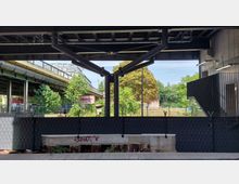 Blick unter eine moderne Brückenkonstruktion mit schwarzen Stahlträgern. Im Hintergrund sind eine begrünte Freifläche, Bäume, ein Geländer mit Graffiti, sowie eine erhöhte Straße mit Rohrleitungen erkennbar.