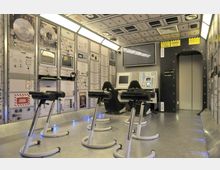 Eine Nachbildung eines Raumes aus einer Raumstation mit technischen Bedienfeldern, Bildschirmen und Kontrollgeräten an den Wänden. Im Vordergrund befinden sich mehrere Trainingssitze und eine glänzende Metallbodenfläche, die mit blauen LED-Lichtern beleuchtet ist.