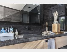 Modernes Badezimmer mit schwarzen Marmorwänden und einer Arbeitsplatte aus dunkelgrauem Stein. Auf der Arbeitsplatte stehen Pflegeprodukte und Dekorationselemente, darunter eine kleine Steinfigur in einer Glasvitrine; darunter befindet sich ein Waschbecken und ein Unterschrank aus hellem Holz.