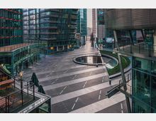 Moderner urbaner Platz, umgeben von Glas- und Stahlarchitektur großer Gebäude. Zentral befindet sich ein kleiner, runder Brunnen, flankiert von bepflanzten Grünflächen und Gehwegen mit diagonalen Markierungen.