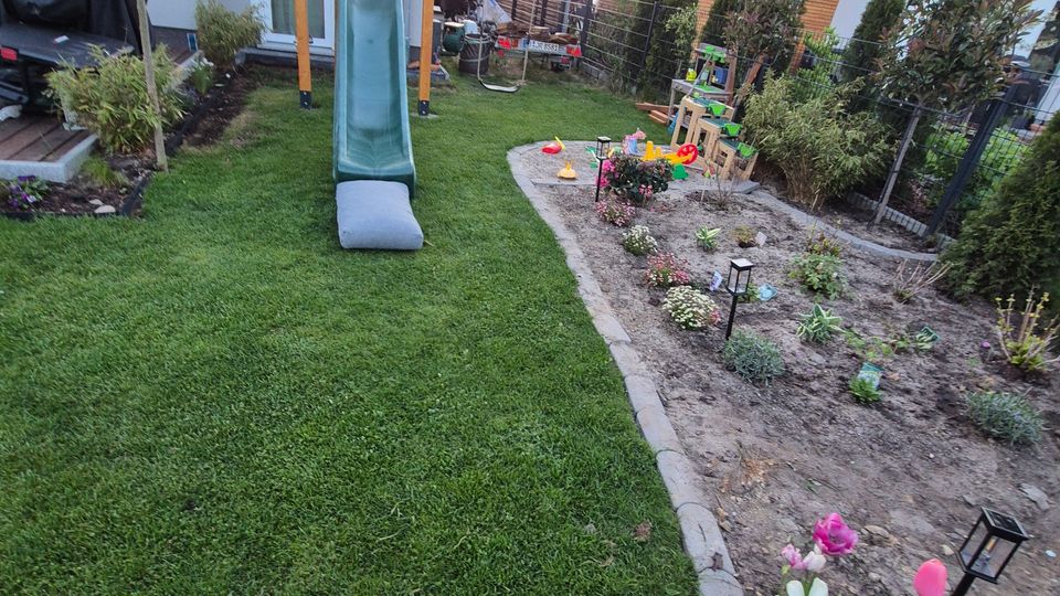 Schmaler Wohnhausgarten mit gepflegtem Rasen und einer Holz-Kletterkonstruktion mit grüner Rutsche in der Mitte. Rechts verläuft ein eingefasstes Blumen- und Beetareal mit kleinen Gartenleuchten und Büschen entlang eines Zauns, umgeben von modernen Wohnhäusern.