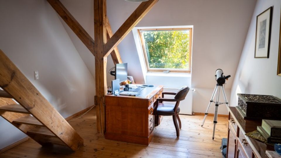 Dachgeschoss-Arbeitszimmer mit sichtbaren Holzbalken und Holzdielenboden, einem Schreibtisch mit Computer sowie einem Stuhl. Ein Dachfenster bringt Tageslicht herein; daneben steht ein Stativ, und an der rechten Wand befinden sich Möbel und gerahmte Bilder.