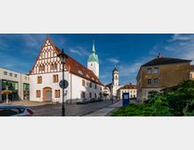 Straßenszene mit einer gotischen Kirche mit weißer Fassade und roten Backsteinverzierungen im Vordergrund, neben modernen und historischen Gebäuden. Im Hintergrund ist ein Kirchturm mit grünem Spitzdach und ein weiterer Turm zu sehen, die Umgebung ist von Kopfsteinpflasterstraßen und geparkten Autos geprägt.