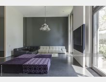 Modernes Wohnzimmer mit grauer Wand, großer Ecksofa-Kombination in Schwarz und Weiß sowie lila gepolsterten Couchtischen mit Kreismuster. Rechts befinden sich bodentiefe Fenster, die den Raum mit Tageslicht erhellen, sowie ein großer, wandmontierter Bildschirm.