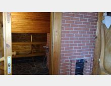 Eingang zu einer Holzsauna mit Holzbänken, Wänden aus Holzpaneelen und einem Boden aus dunklen Steinfliesen. Rechts ist eine Wand aus roten Ziegelsteinen mit einem kleinen Ofenfach sichtbar.