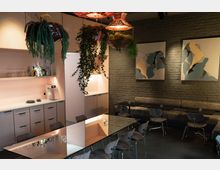 Ein modernes Café mit einer minimalistisch eingerichteten Bar im Hintergrund, hellrosa Schränken und Regalen mit Gläsern und Getränkeflaschen. Der Sitzbereich hat gepolsterte Bänke, dunkle Holztische, graue Stühle und eine Ziegelwand mit abstrakten Wandgemälden, während von der Decke hängende Pflanzen den Raum dekorieren.