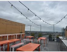 Rooftop terrace dining area with orange tables, striped benches, and metal chairs beneath string lights, bordered by railings and planter boxes. The terrace overlooks a wide city skyline under an overcast sky, with a wood-paneled wall on one side.