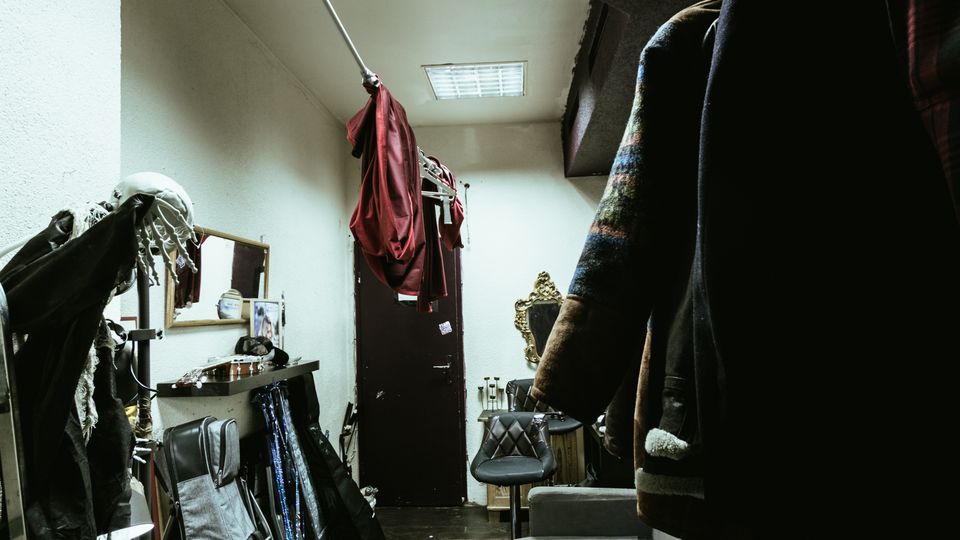 Narrow backstage dressing room or storage corridor with fluorescent ceiling lights, wood plank flooring, and white walls. Clothing and coats hang along the right side, while instrument cases, a mirror and shelf, and scattered gear line the left, leading to a door at the far end.