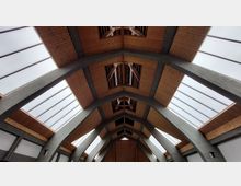 Dachansicht eines modernen Gebäudes mit einer Holzkonstruktion und großen, schrägen Glasflächen, die von dunkelgrauen Stahlträgern gestützt werden. Die Gestaltung zeigt eine symmetrische Anordnung mit geometrischen Formen und natürlichem Licht.