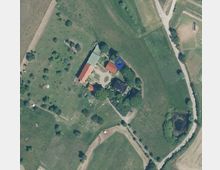 Luftaufnahme eines ländlichen Anwesens mit mehreren Gebäuden, darunter zwei rote Ziegeldachhäuser und eine große grüne Freifläche. Umgeben von Wiesen, Bäumen und einem kleinen Teich, mit Wegen, die das Gelände verbinden.