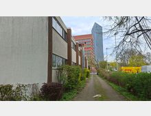 Schmaler gepflasterter Weg zwischen niedrigen Wohn- oder Bürogebäuden mit flachen Dächern und Sträuchern am Rand. Im Hintergrund ragen ein rotes Hochhaus und ein modernes Glashochhaus über Bäume und Straßenlampen hinweg.