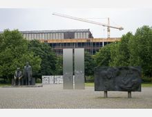 Platz mit einer Karl-Marx- und Friedrich-Engels-Statue, flankiert von einer großen Metallskulptur und einem Relief mit menschlichen Figuren, umgeben von Bäumen. Im Hintergrund ist eine Baustelle mit einem gelben Baukran und einem teilsanierten Gebäude zu sehen.
