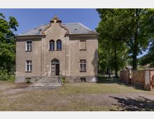 Zweistöckiges, historisches Haus mit Putzfassade und geschwungenem Giebel, zentraler Eingangstür mit Treppe, umgeben von Bäumen und einer kleinen gemauerten Gartenmauer auf der rechten Seite.