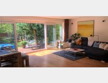 Helles Wohnzimmer mit Holzfußboden, einer großen Eckcouch, Couchtisch und Dekoration. Eine Glas-Schiebetür führt auf eine Terrasse mit Pflanzen und Blick in einen Garten mit Bäumen und Gartenmöbeln.