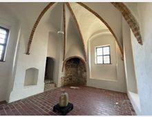 Innenraum eines historischen Gebäudes mit spitzbogigen Gewölbedecken aus Ziegeln, weiß verputzten Wänden und einem Boden aus roten Backsteinen. Zwei Fenster lassen Tageslicht herein, und in der Mitte des Raumes steht ein kleiner Stein auf einem schwarzen Sockel.