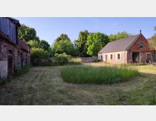 Ein ländlicher Innenhof mit zwei Gebäuden aus Ziegel- und Naturstein, umgeben von Bäumen und Büschen. Im Vordergrund befindet sich eine Wiese mit gemähtem und hohem Gras.