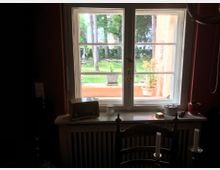 Blick aus einem Fenster in einem dunklen Raum auf einen Garten mit grünem Rasen, Bäumen und Außenmöbeln. Auf der Fensterbank stehen verschiedene Gegenstände, darunter ein Retro-Radio und eine Pflanze, und im Vordergrund ist eine Stuhllehne zu sehen.