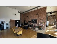 Offener Wohnraum in einem industriellen Loft-Stil mit sichtbaren Backsteinwänden, hohen Decken und großen Fenstern. Der Raum verfügt über eine Sitzecke mit Sofa, eine Kücheninsel mit Barhockern, einen Esstisch mit gelben Stühlen und hellem Holzboden.