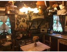 Rustikale Küche mit Holzdecken und nostalgischer Einrichtung. Kupfertöpfe und Laternen hängen von der Decke, während Regale, ein alter Herd und ein Tisch mit Spitzentischdecke den Raum prägen; durch die Fenster mit Spitzenvorhängen ist grüne Vegetation sichtbar.