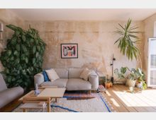 Wohnzimmer mit einer grauen Couch, einem kleinen Holz-Couchtisch und großen Zimmerpflanzen. Die Wände sind unbehandelt und teilweise rau, während Tageslicht durch eine geöffnete Balkontür einfällt. Ein buntes abstraktes Bild hängt an der Wand.