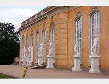 Seitenansicht eines historischen Gebäudes mit ockerfarbener Fassade und hohen weißen Fenstern im barocken Stil. Vor dem Gebäude stehen weiße Marmorstatuen auf Sockeln entlang eines Kieswegs, umgeben von Bäumen im Hintergrund.