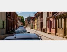 Gepflasterte Straße in einer historischen Altstadt mit schmalen, dicht beieinander stehenden Häusern in verschiedenen Pastelltönen und Backsteinfassaden. Entlang der Straße sind parkende Autos und einige Fensterläden an den Gebäuden geöffnet, während im Hintergrund Bäume sichtbar sind.