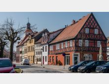 Straßenszene in einem historischen Stadtzentrum mit Fachwerkhäusern in verschiedenen Farben, darunter ein auffälliges rotes Gebäude. Die gepflasterte Straße wird von geparkten Autos und kahlen Bäumen gesäumt, im Hintergrund ist ein Kirchturm mit Wetterfahne zu sehen.