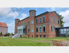 Historisches Backsteingebäude mit grünen Fensterrahmen und Türmchen an den Ecken, ergänzt durch einen modernen Glasanbau auf der rechten Seite; im Vordergrund eine gepflegte Rasenfläche.