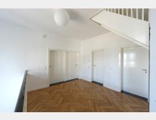 Flur in einem Wohngebäude mit hellem Fischgrätenparkettboden, weißen Wänden und vier weißen Zimmertüren sowie einer offenen Treppe rechts oben im Bild. Eine kugelförmige Deckenleuchte ergänzt das minimalistische Design.