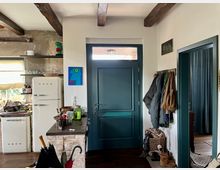 Eingangsbereich in einem gemütlichen Wohnhaus mit Holzbalkendecke, blauer Haustür, Garderobe mit Jacken und Körben sowie angrenzender Küche. Links ein Retro-Kühlschrank, offene Regale und eine helle Küchentheke mit Utensilien.