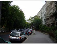 Straßenansicht in einer städtischen Wohngegend mit mehrstöckigen historischen Gebäuden mit verzierten Fassaden auf der rechten Seite und einer Baumreihe auf der linken Seite entlang der Straße. Geparkte Autos und Motorräder säumen die Straße und den Gehweg.