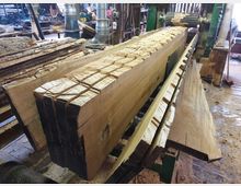 Ein Sägewerk-Innenraum mit einem großen Holzblock auf einer Maschine, der teilweise zugesägt ist. Umgeben von Holzteilen, Spänen und Maschinenteilen im Hintergrund.