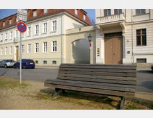 Eine Straßenszene mit einem historischen Gebäude mit hellen Fassaden und Sprossenfenstern im Hintergrund. Im Vordergrund steht eine große Holzbank, daneben ein Parkverbotsschild, und auf der Straße parken einige Autos.