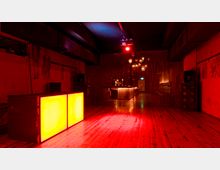 Ein dunkler Clubraum mit Holzboden, roten Wänden und gedämpfter Beleuchtung. Im Vordergrund steht ein leuchtendes DJ-Pult, im Hintergrund befindet sich eine Bar mit Lichtern, die durch einen Vorhang aus dünnen Fäden teilweise verdeckt ist.