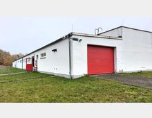 Flaches Gebäude mit weißer Fassade, großem roten Rolltor und angrenzender roter Eingangstür, umgeben von einer Wiese. Im Hintergrund sind Bäume in herbstlichen Farben sichtbar.