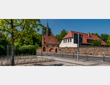 Historisches Gebäudeensemble mit dem St. Georgen-Hospital, bestehend aus einem Fachwerkgebäude mit rotem Ziegeldach und einem mittelalterlichen Torhaus mit Turmspitze. Im Vordergrund eine Pflasterstraße, Fahrradständer und ein Baum, im Hintergrund Grünflächen und Bäume.