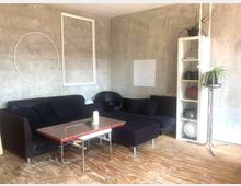 Wohnraum mit modernem, minimalistischem Stil: schwarzes Sofa mit passenden Kissen, ein Tisch mit Granitplatte, freiliegende Betonwände mit weißen Rahmenelementen als Dekoration. Rechts ein hohes Regal mit Dekorationsgegenständen und Pflanzen; Holzbodenbelag sichtbar.