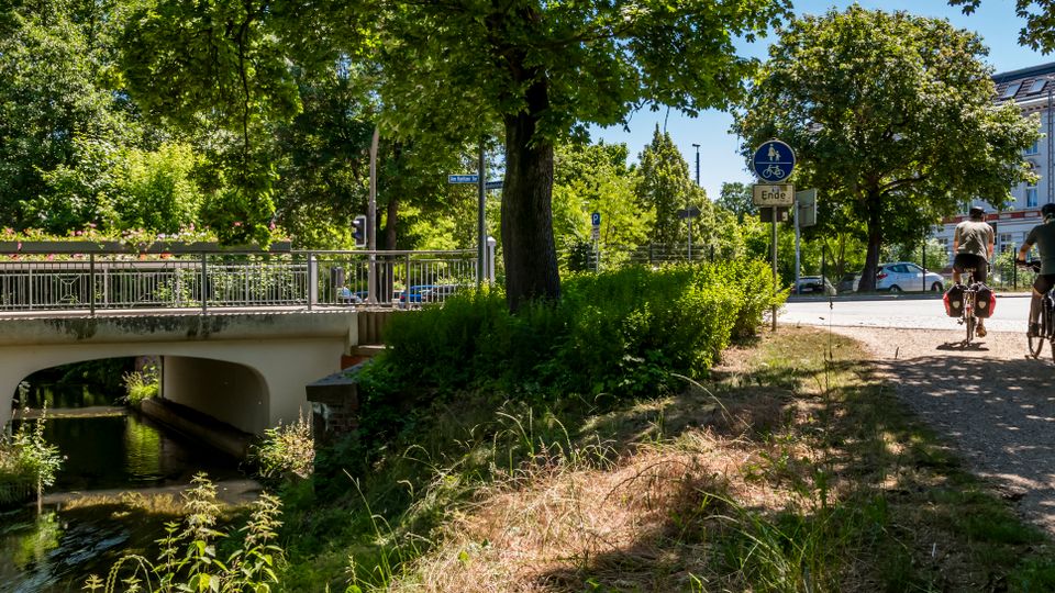 Uferweg an einem kleinen Bach mit einer niedrigen Betonbrücke und Metallgeländer, umgeben von dichtem Grün und großen Bäumen. Rechts verläuft ein Schotterweg neben einer Straße, im Hintergrund steht ein mehrstöckiges, helles Gebäude mit vielen Fenstern.
