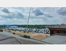 Blick von einem Dach auf eine moderne Wohnsiedlung mit weiß verputzten, zweigeschossigen Reihenhäusern und schwarzen Fensterrahmen. Im Hintergrund ein Gebäude mit blauer Dachkonstruktion und eine grüne Landschaft mit Bäumen und einem Fluss.