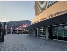 Außenansicht der Mercedes-Benz Arena in Berlin mit einer breiten, gepflasterten Fläche im Vordergrund, geschwungenem Vordach mit dem Arena-Schriftzug rechts und einem modernen Gebäude mit Werbetafeln im Hintergrund.